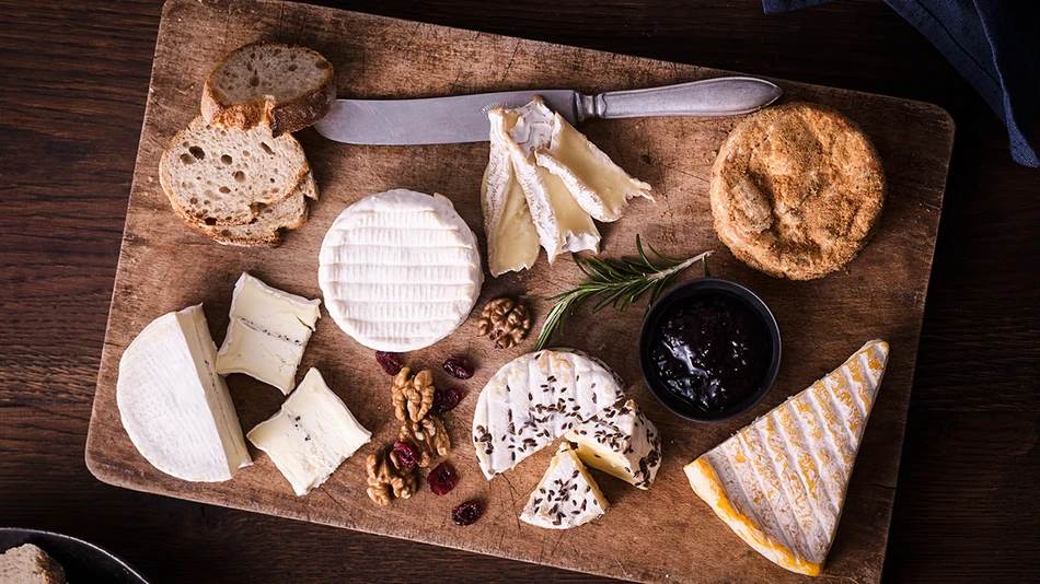 A range of soft cheeses on a wooden board, placed on a wooden table, with a small bowl of fruit preserve, some walnuts and dried
