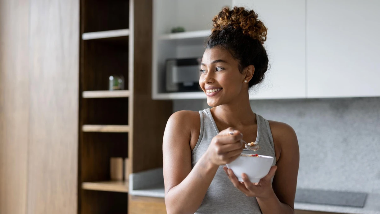 Woman eating yogurt in a kitchen from a white bowl
