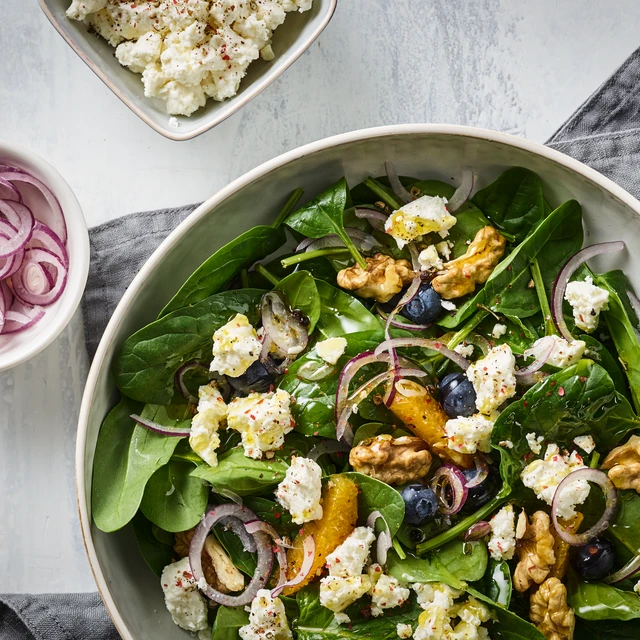 Fresh salad in a large bowl with mixed greens, walnuts, blueberries, orange slices, red onion rings, and crumbled feta cheese. The bowl sits on a light surface with a gray cloth nearby, and two smaller bowls in the corner hold pickled red onions and more crumbled feta cheese.