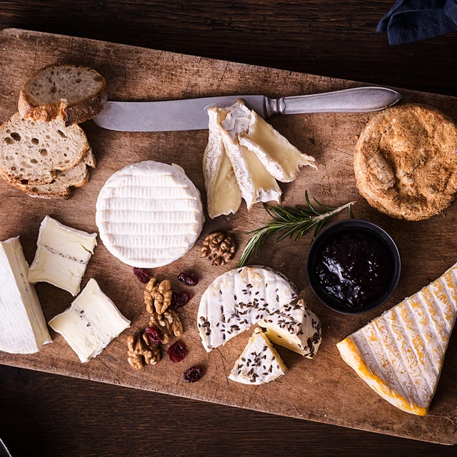 A range of soft cheeses on a wooden board, placed on a wooden table, with a small bowl of fruit preserve, some walnuts and dried