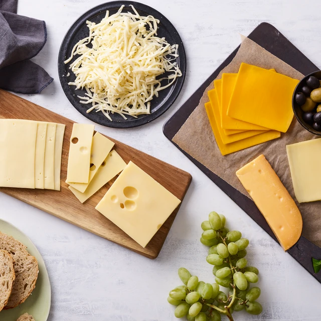 Flat lay de uma seleção de queijos variados em tábuas de madeira e pratos, acompanhados de uvas verdes, azeitonas, pão rústico e folhas verdes, sobre uma superfície clara.