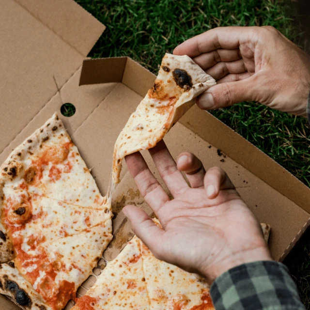 Hands hold a slice of cheese pizza above an open cardboard pizza box placed on grass. Melted cheese stretches between the lifted slice and the remaining pizza, highlighting the pizza’s soft, elastic texture.
