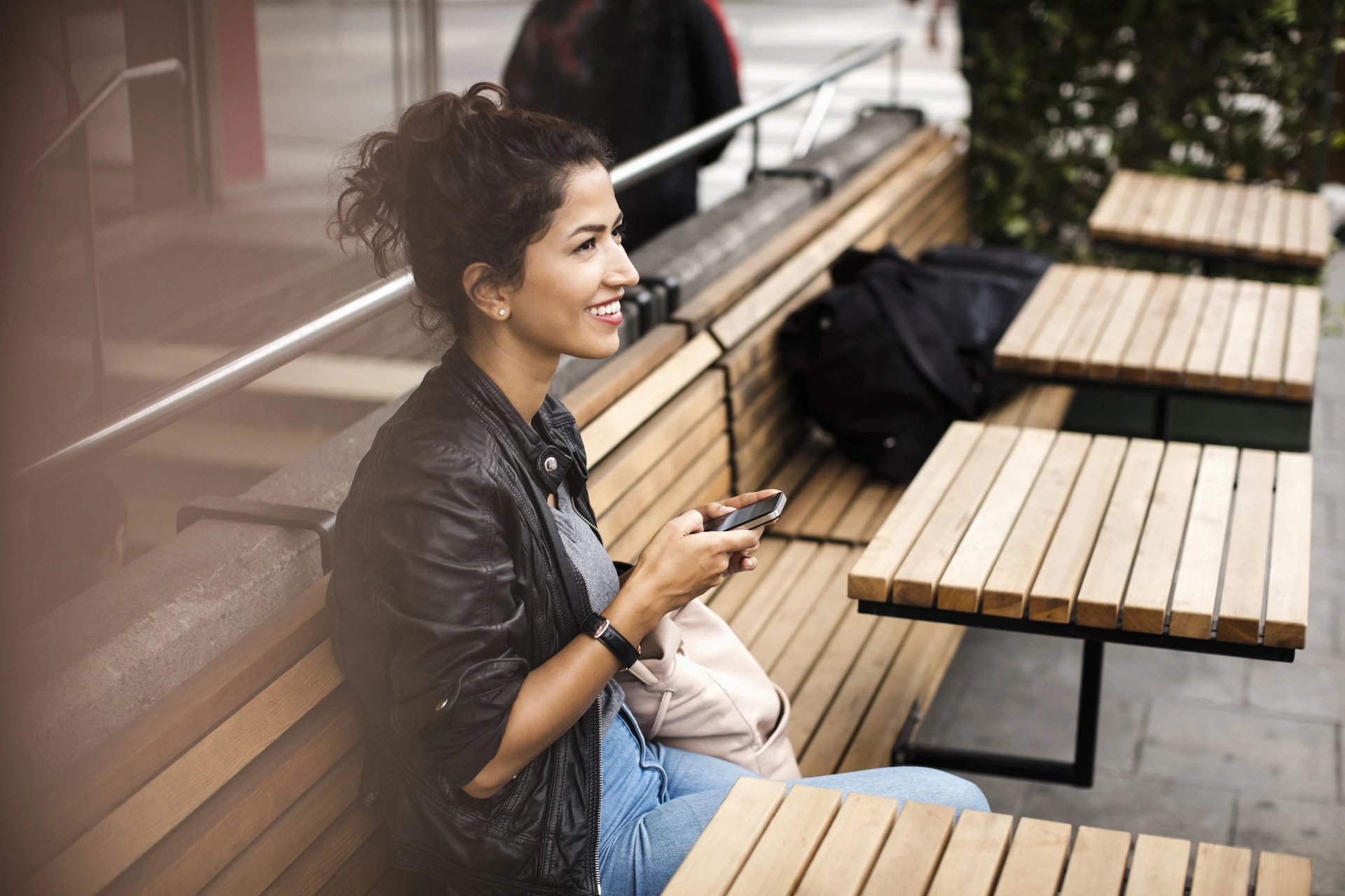 Woman wearing a black leather jacket sitting on a wooden bench with mobile phone on hand