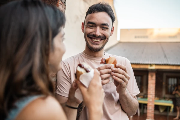 Young man talking and eating choripán with friend in the backyard at home
