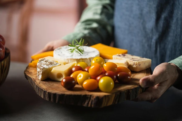 A person in a blue apron holding a wooden cheeseboard with various cheeses and cherry tomatoes 