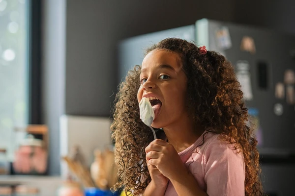 Girl with curly hair in the kitchen, licking a spoon covered in a dairy product 