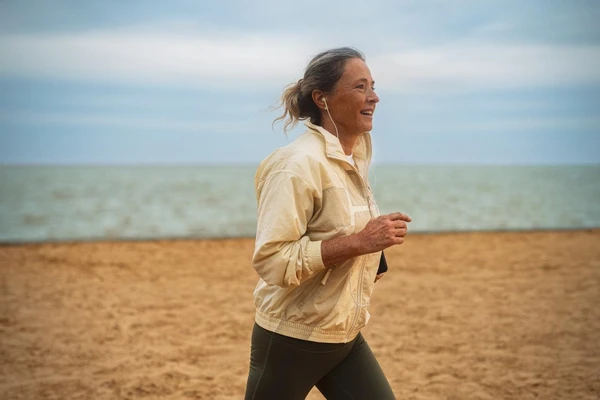Older woman wearing headphones listening to music while running on beach. 