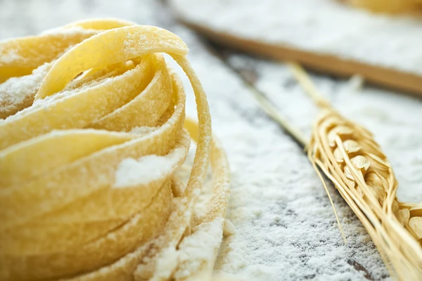 Uncooked pasta nests dusted with flour on a floured surface, with wheat stalks in the background.