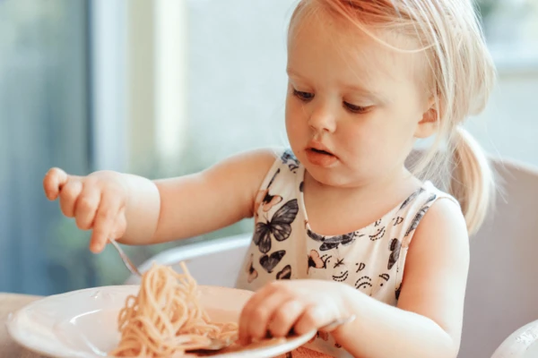 A young child sits at a wooden table, using one hand to pick up noodles from a white bowl while the other hand rests near the food.