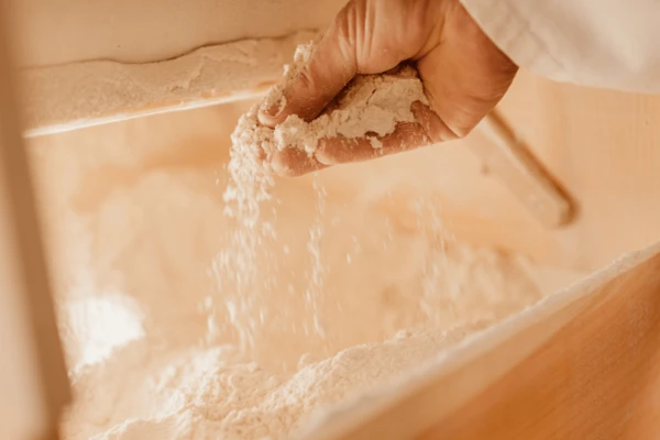 A hand sprinkles white flour into a wooden container filled with flour.