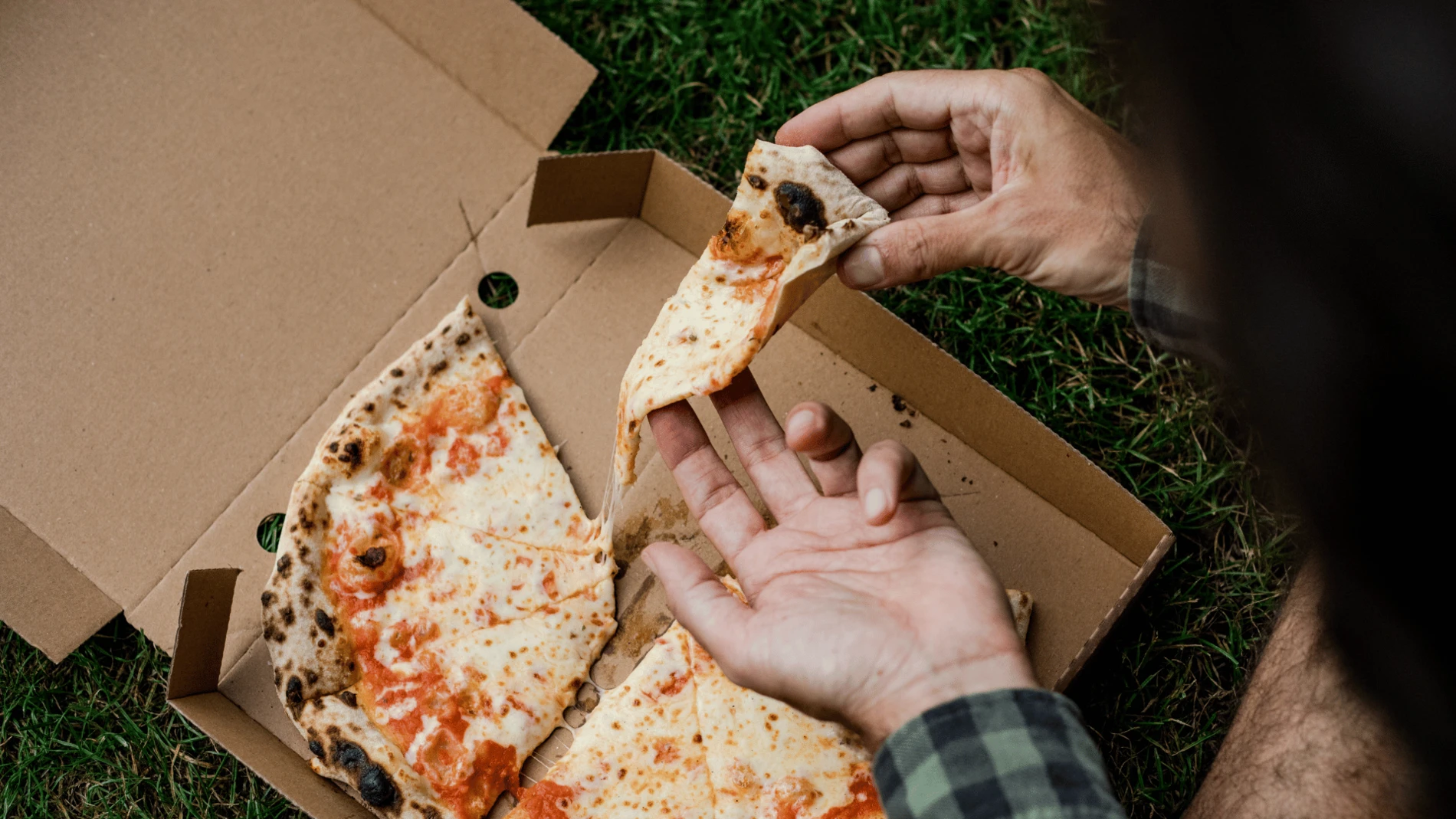 Hands hold a slice of cheese pizza above an open cardboard pizza box placed on grass. Melted cheese stretches between the lifted slice and the remaining pizza, highlighting the pizza’s soft, elastic texture.