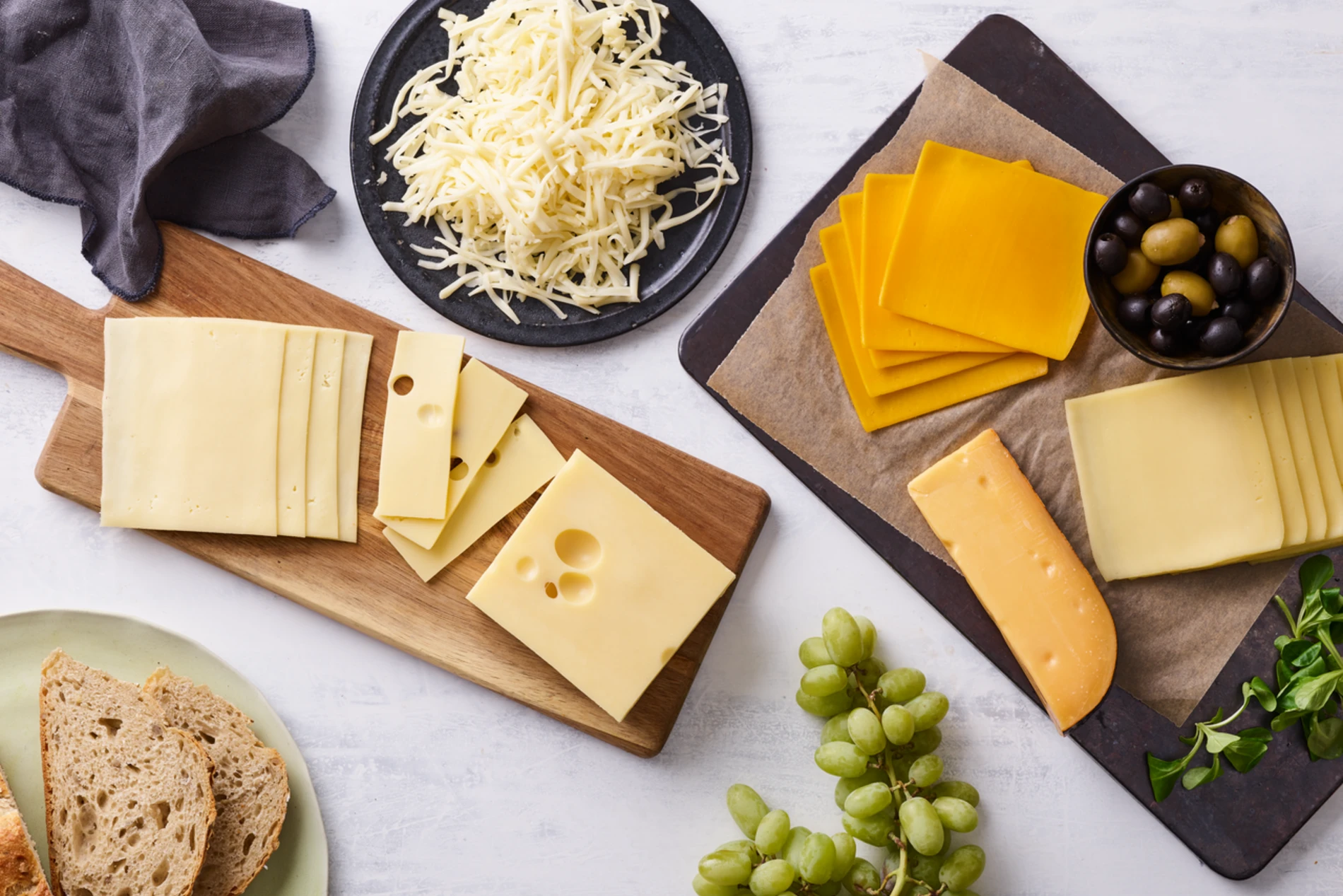 Flat lay de uma seleção de queijos variados em tábuas de madeira e pratos, acompanhados de uvas verdes, azeitonas, pão rústico e folhas verdes, sobre uma superfície clara.
